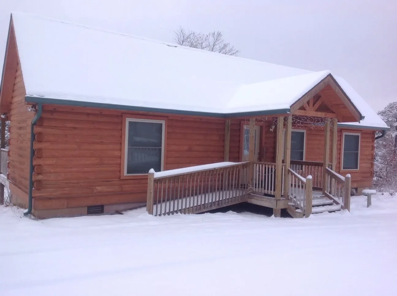 A wooden house with a snow-covered roof and a small front porch in a snowy landscape.