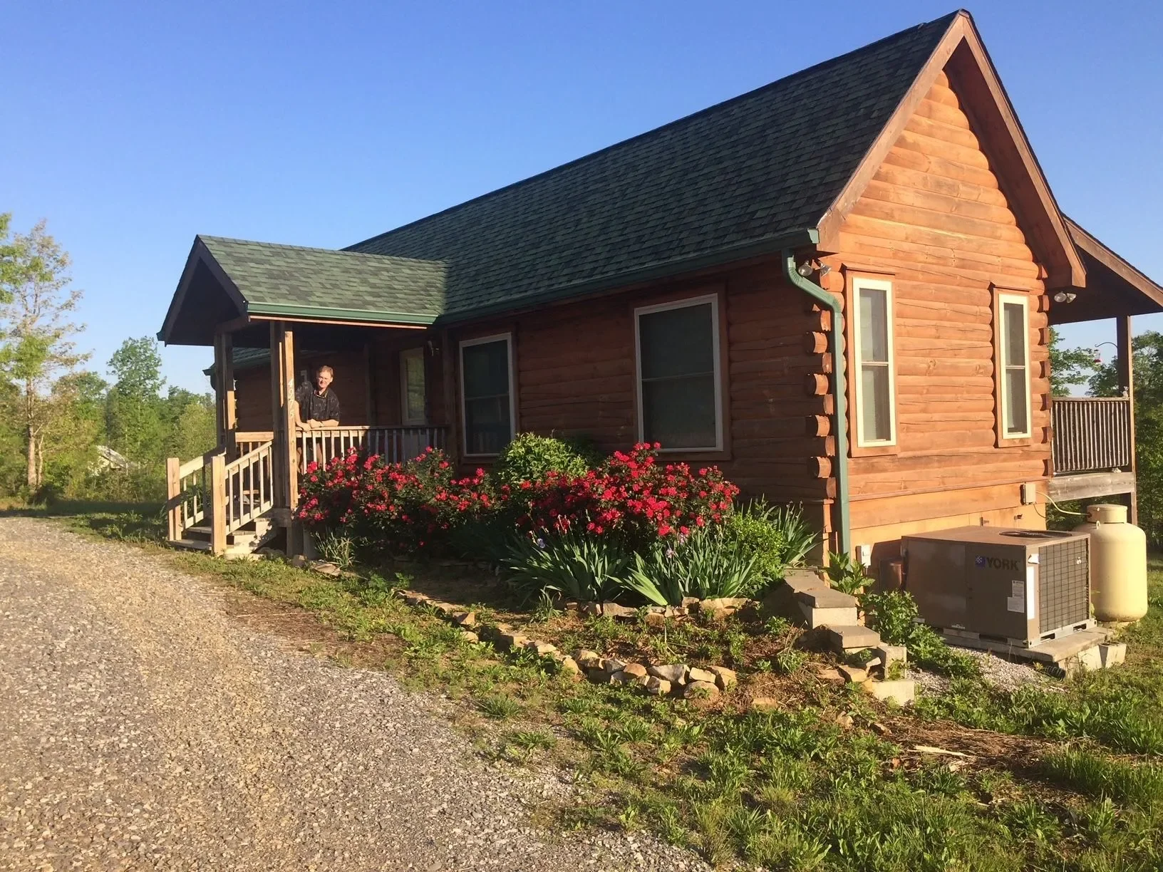 A wooden house with a porch, three windows, and a garden with pink flowers. A person is standing on the porch, looking outside. The house is surrounded by trees and a gravel path.