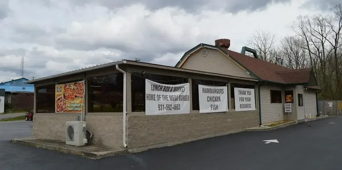 Corner view of a small fast-food restaurant with signs advertising pizza, burgers, chicken, fish, and business takeout, located next to parking lot and trees under cloudy sky.