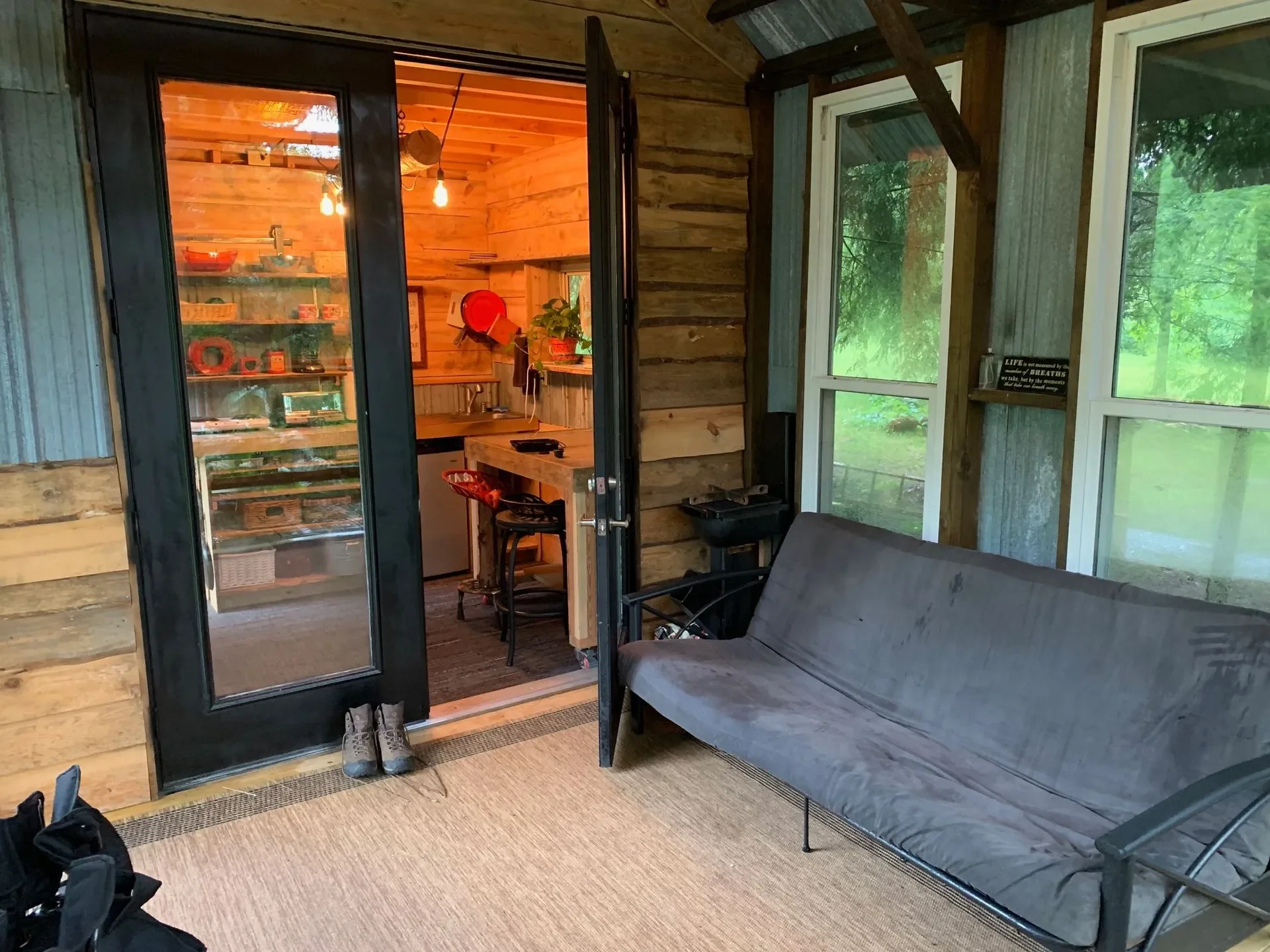 Indoor porch area with a gray futon, three windows showing greenery outside, and a view into a wood-paneled kitchen with open shelves, a sink, and hanging lights.