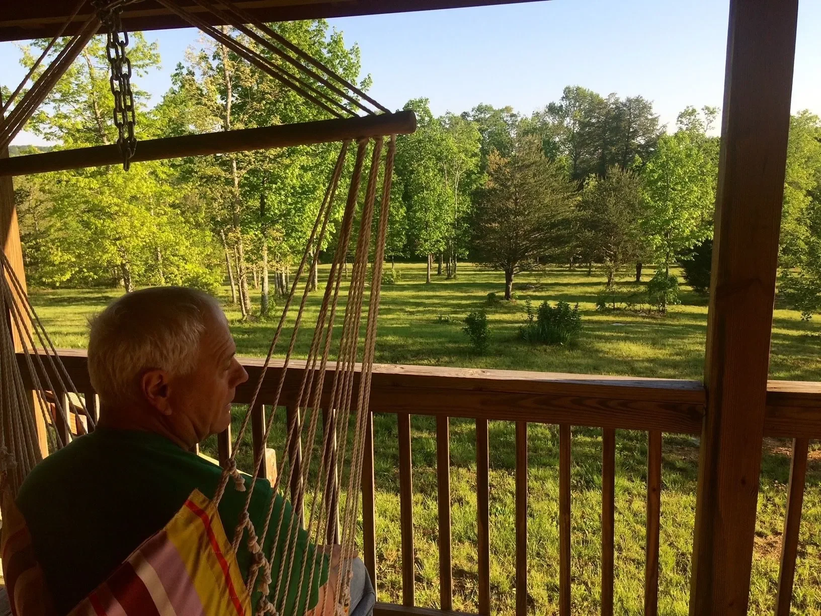 An elderly man sitting on a porch swing, overlooking a lush green lawn and trees in the distance, during daylight.