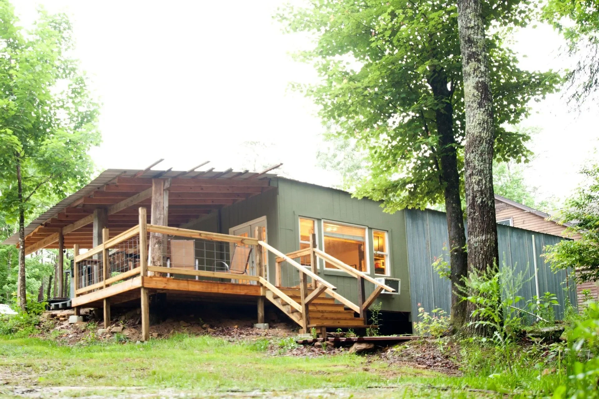 A raised, green, metal-sided cabin with large windows, a wooden porch, and a set of stairs, surrounded by trees and greenery.