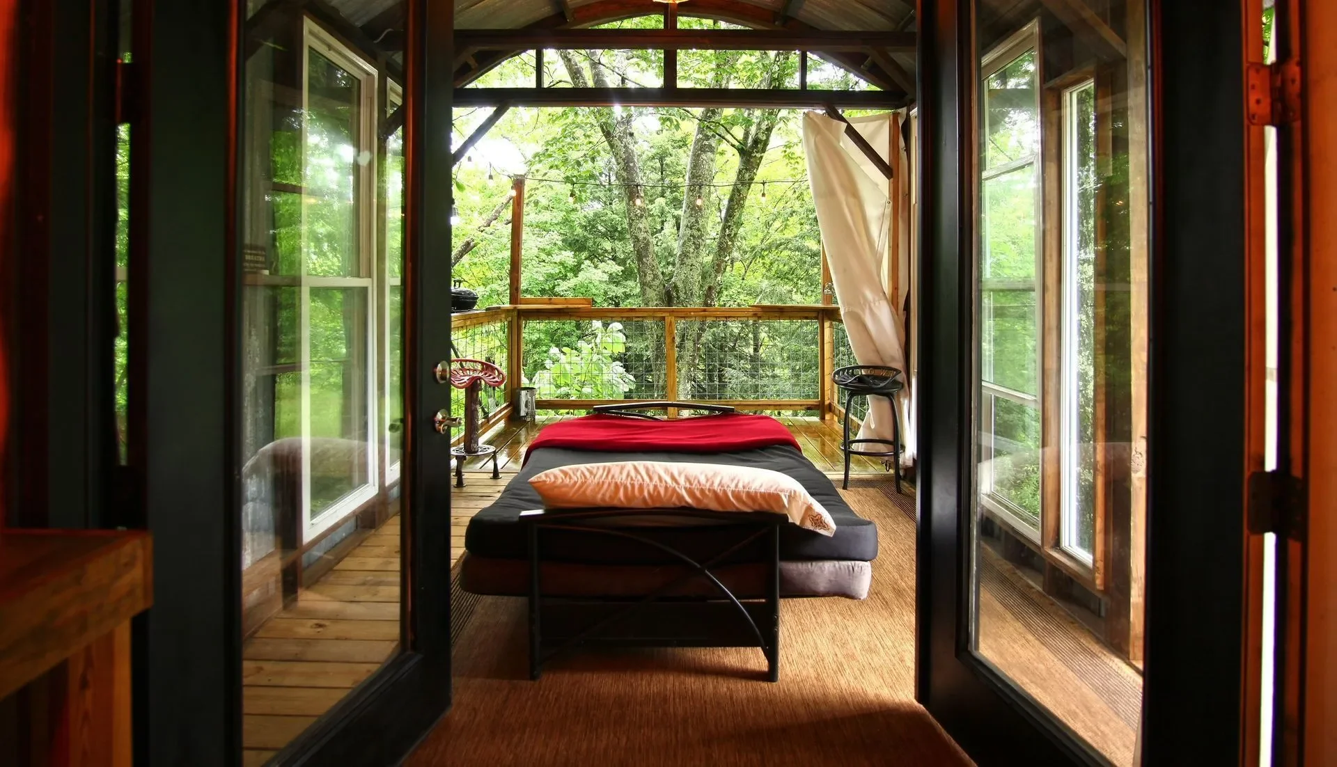 A cozy bedroom with a bed and a view of a green forest outside, seen through open wooden framed glass double doors.
