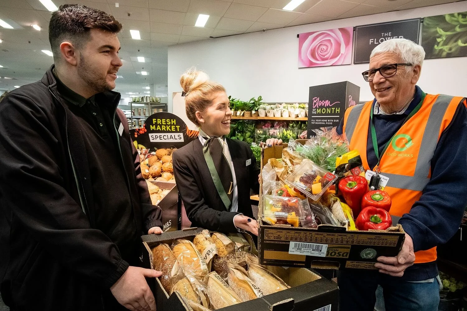 Two smiling M&S employees holding a donation box of M&S food items, with a community foodbank employee also holding the donations box.