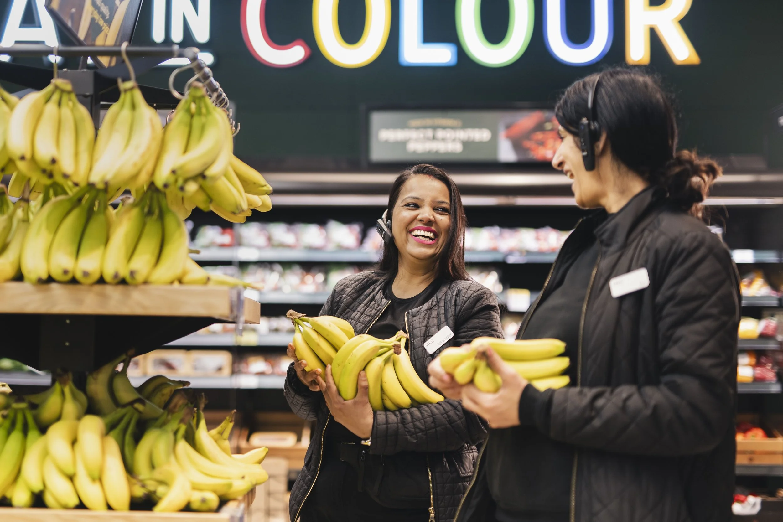 Two M&S employees restocking bananas in the produce section, smiling and talking.