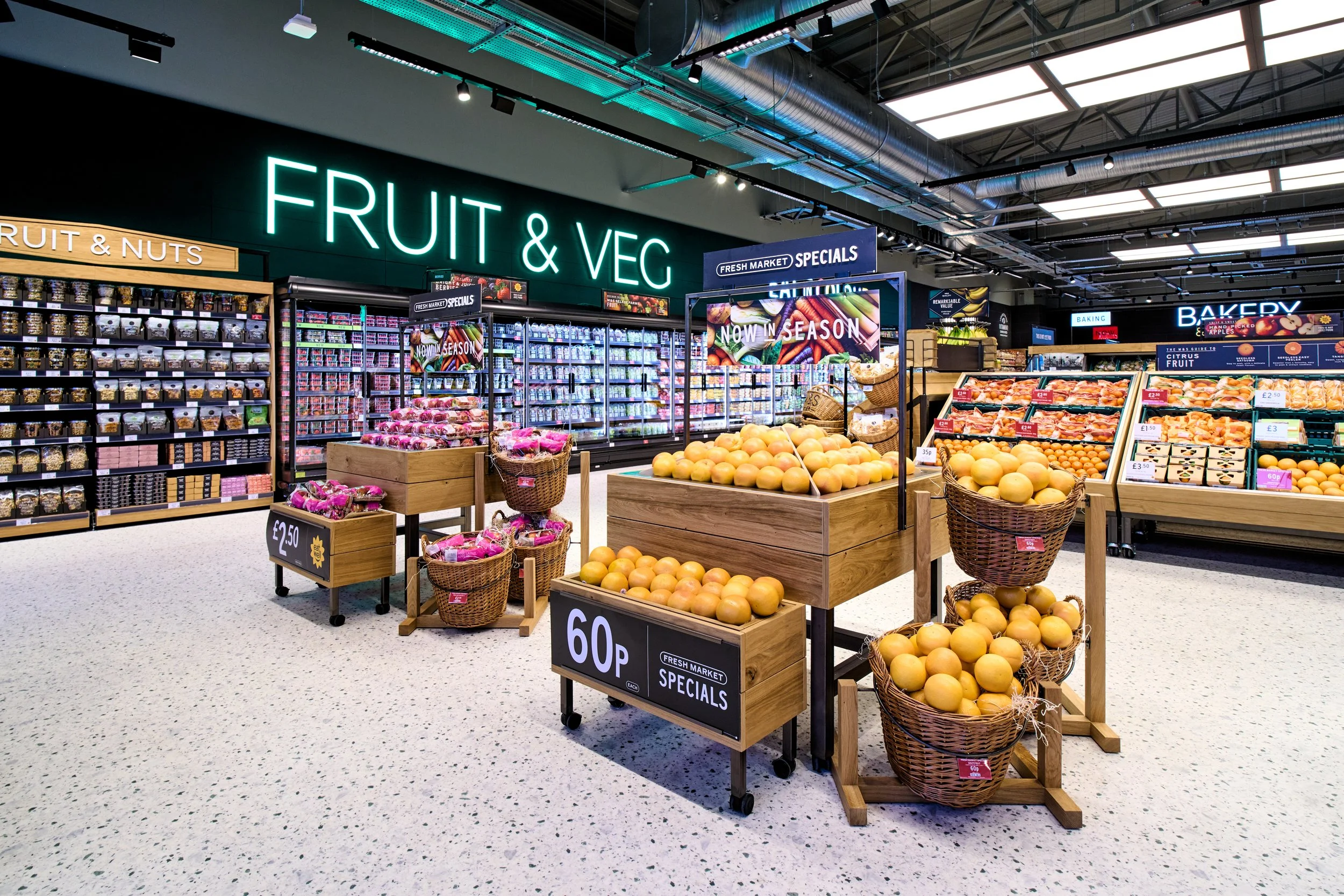 M&S produce section with fruits and vegetables, including bananas, oranges, and peaches, displayed in wooden bins and baskets, with neon signs indicating sections for fruit andvegetables.