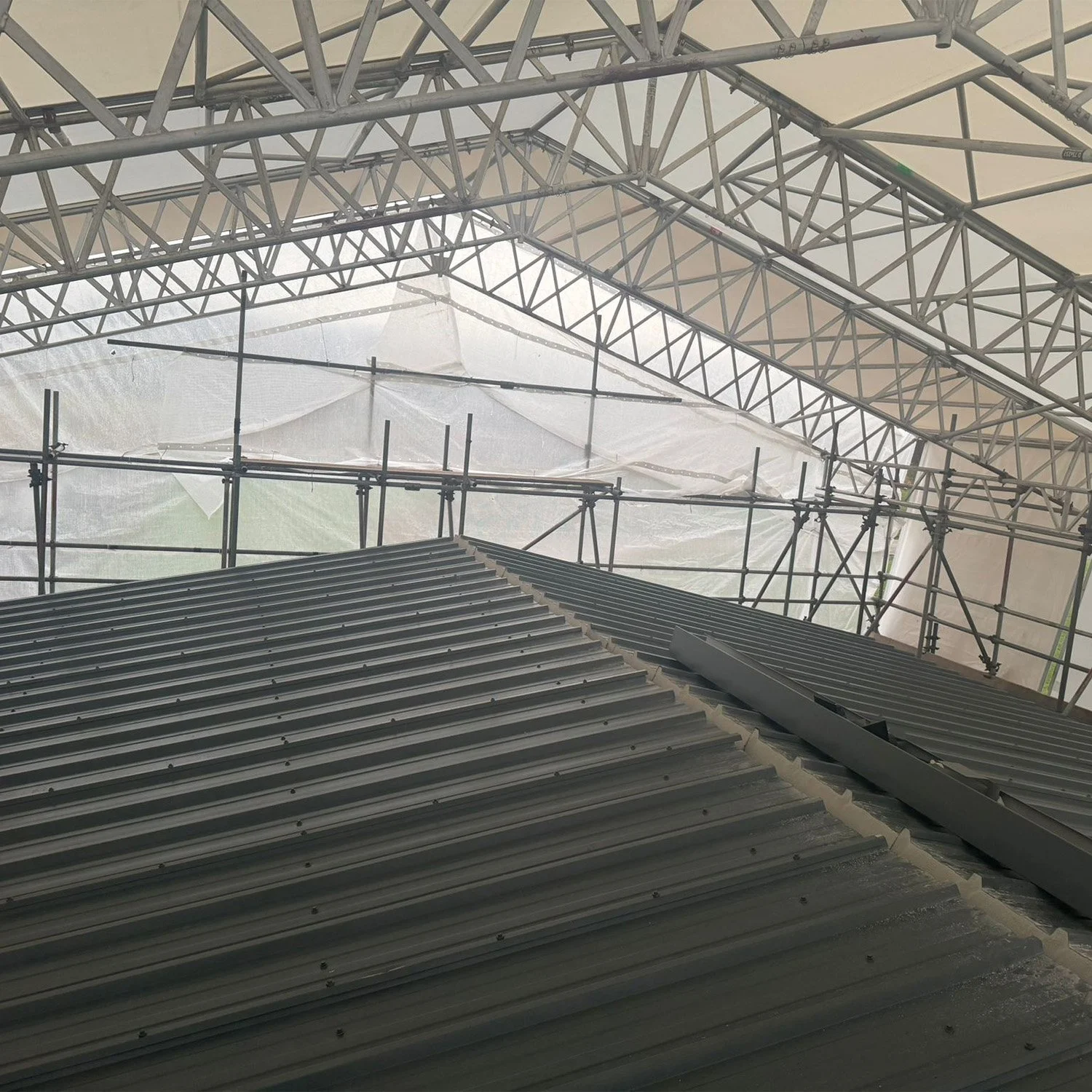 Construction site with metal roofing panels and scaffolding inside a large tent or temporary structure.