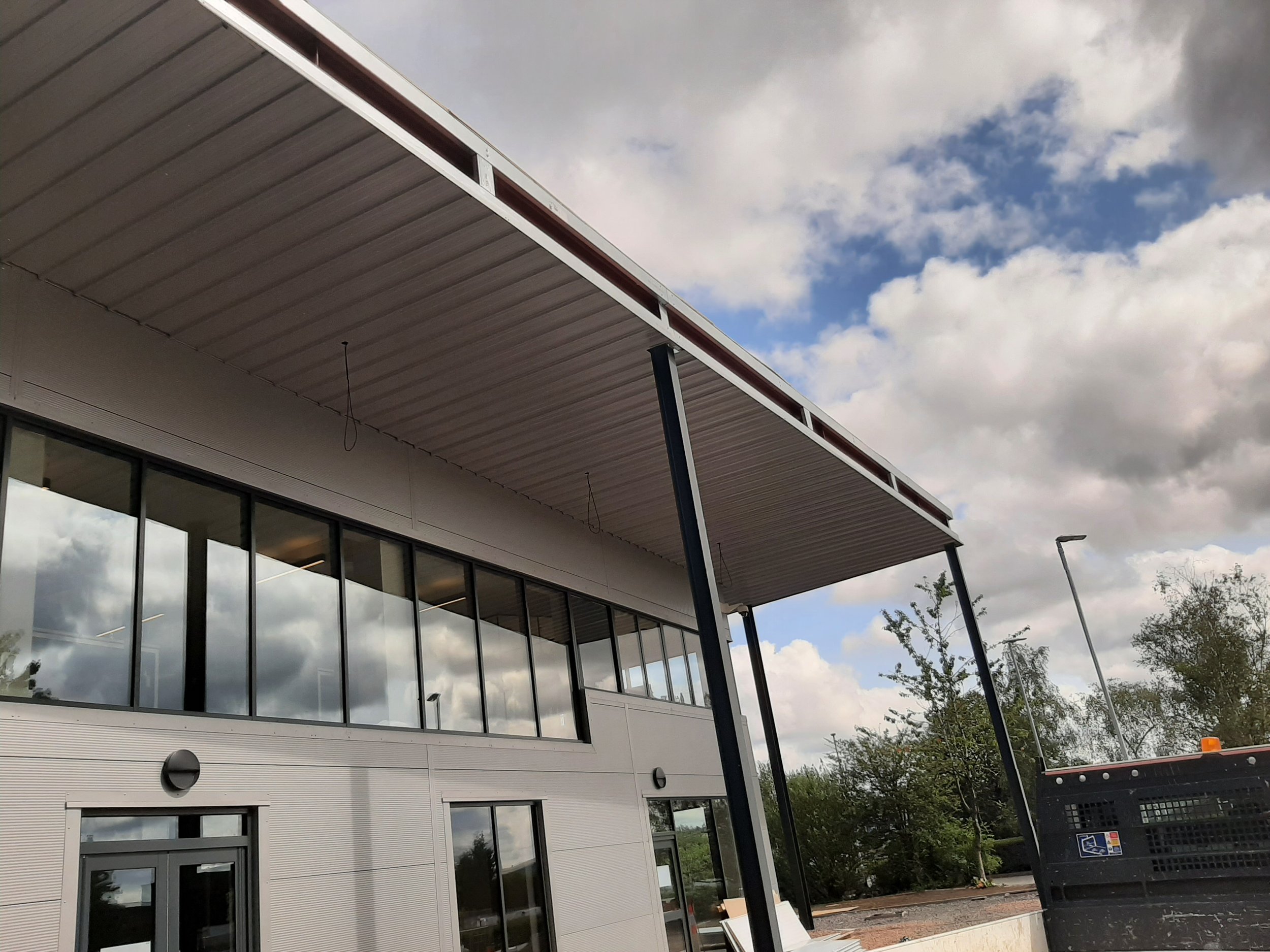 Exterior view of a modern building with large glass windows and metal siding, featuring an overhanging roof and supporting columns, under a partly cloudy sky.