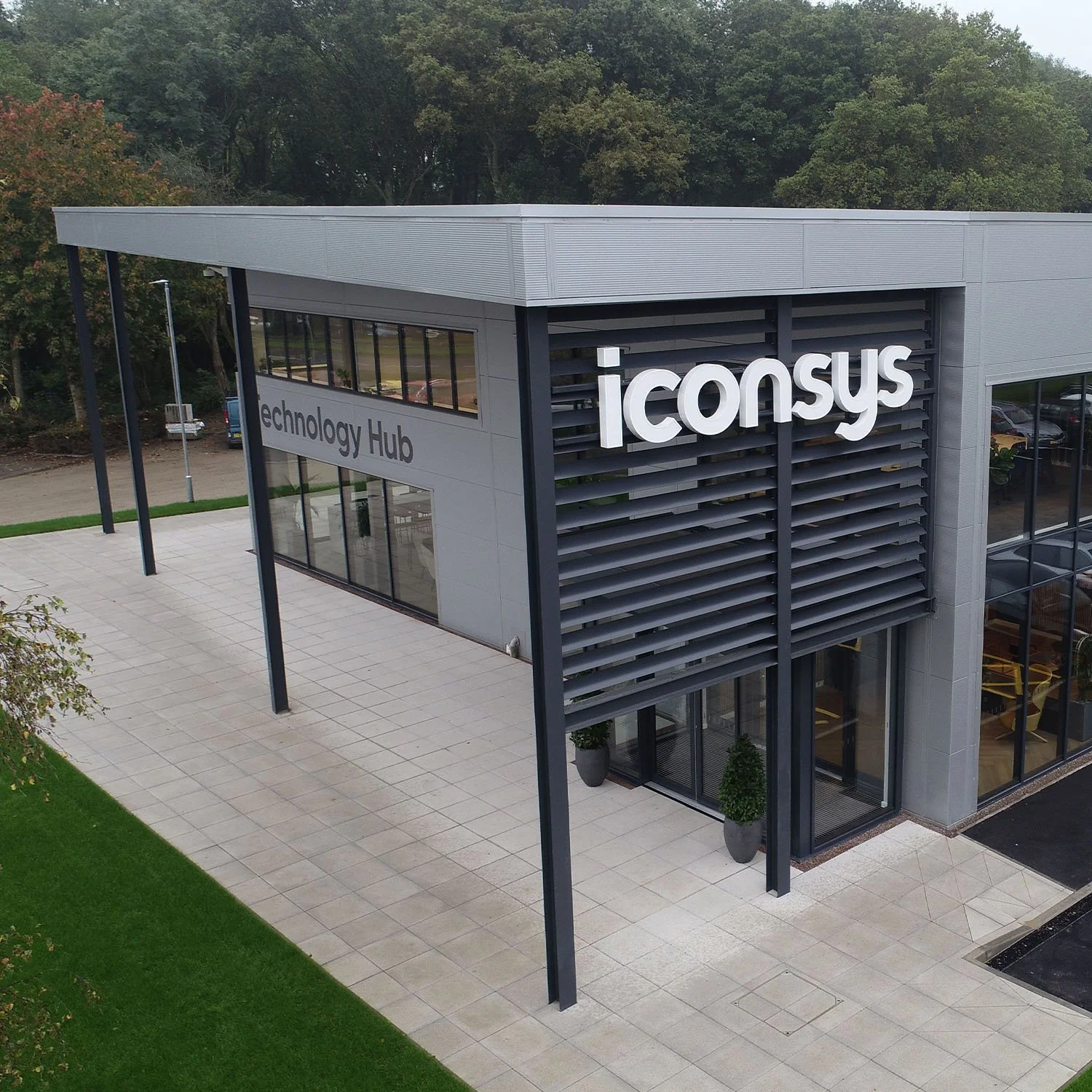 Modern two-story building labeled 'iConsys' with large windows, labeled 'Technology Hub', surrounded by greenery, including trees and grass, with a tiled walkway in front.