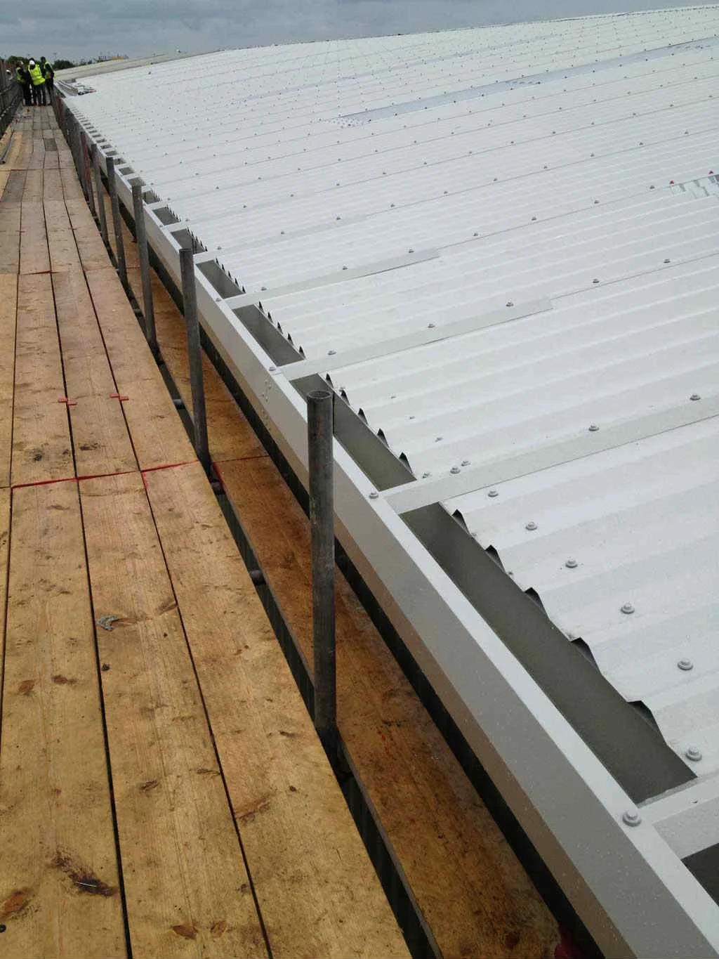 Construction workers installing a white metal roof on a building with a wooden scaffolding walkway.