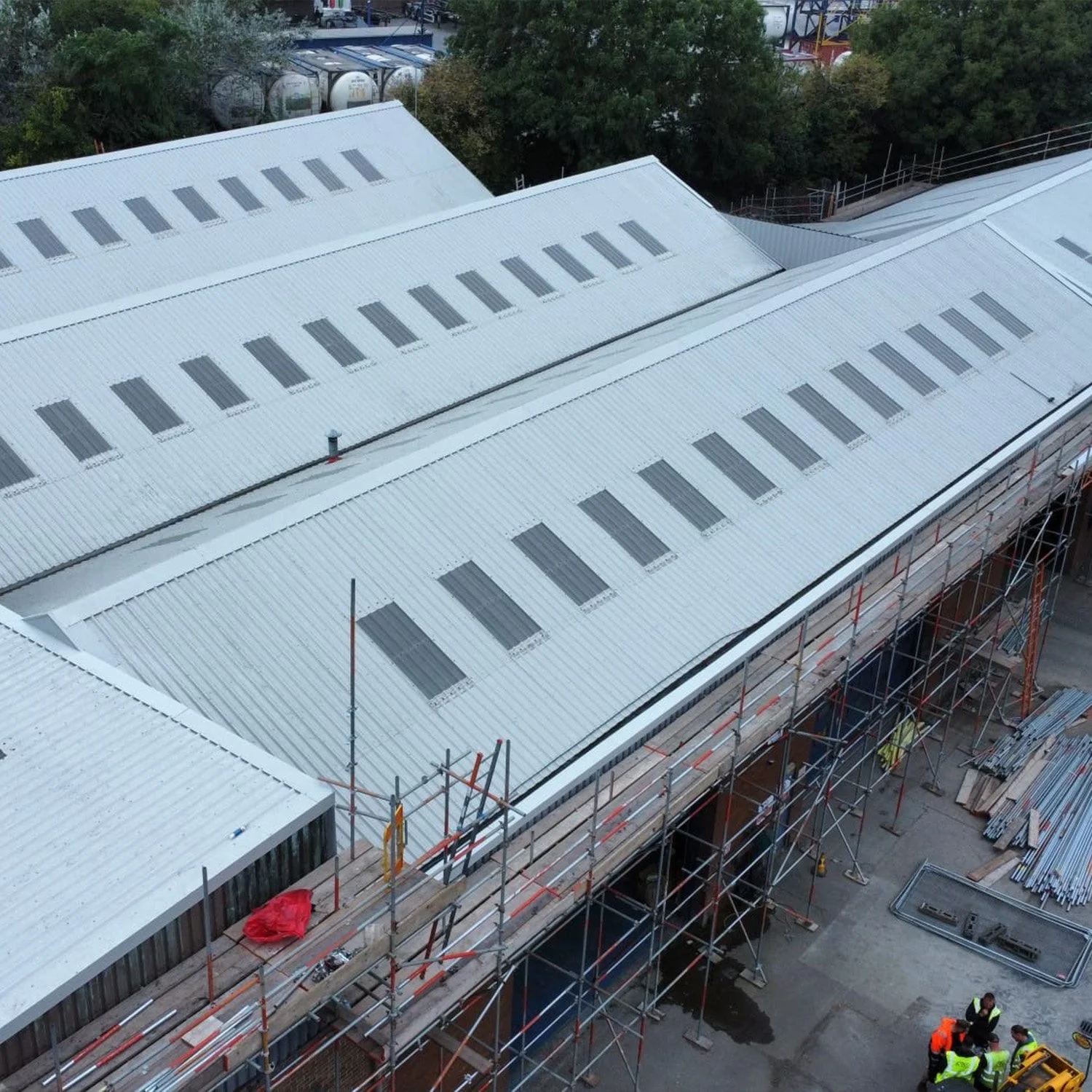 Construction site with metal roofing and scaffolding, workers in safety vests, and covered materials at the site.