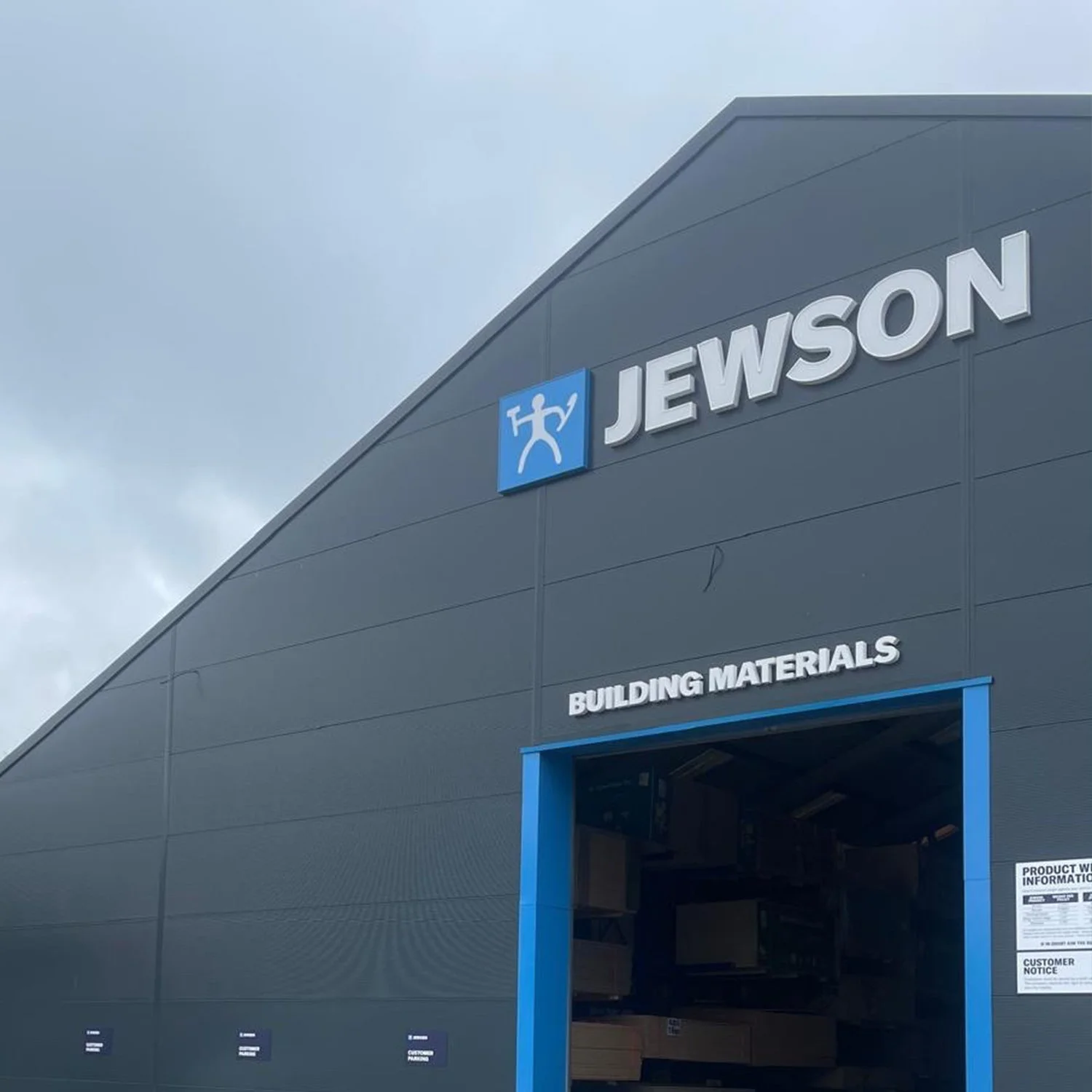 Exterior view of a building materials store called Jewson with a blue sign and logo, grey walls, and a partially open entrance.