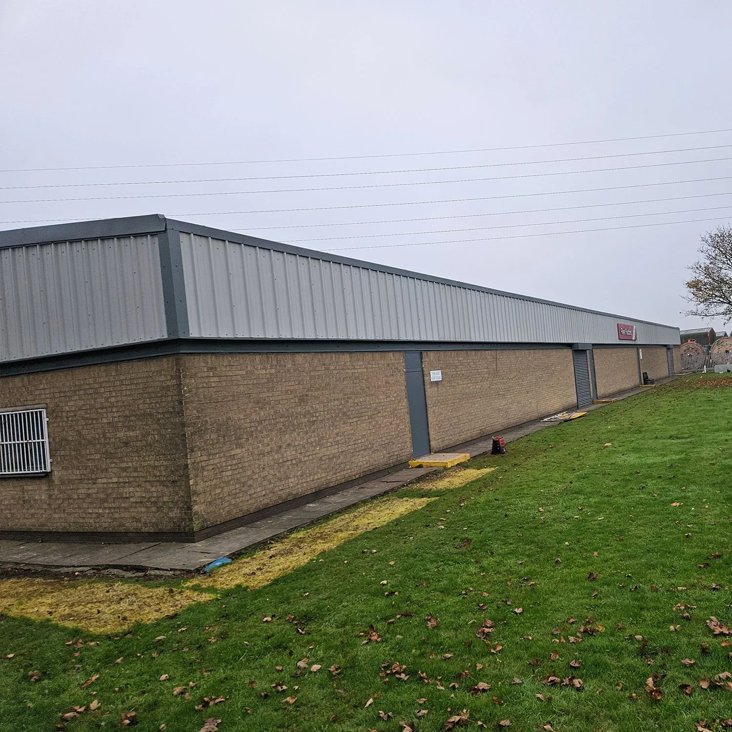 A large rectangular building with brick walls and a gray corrugated metal roof, situated on a grassy area with fallen leaves, under a cloudy sky.