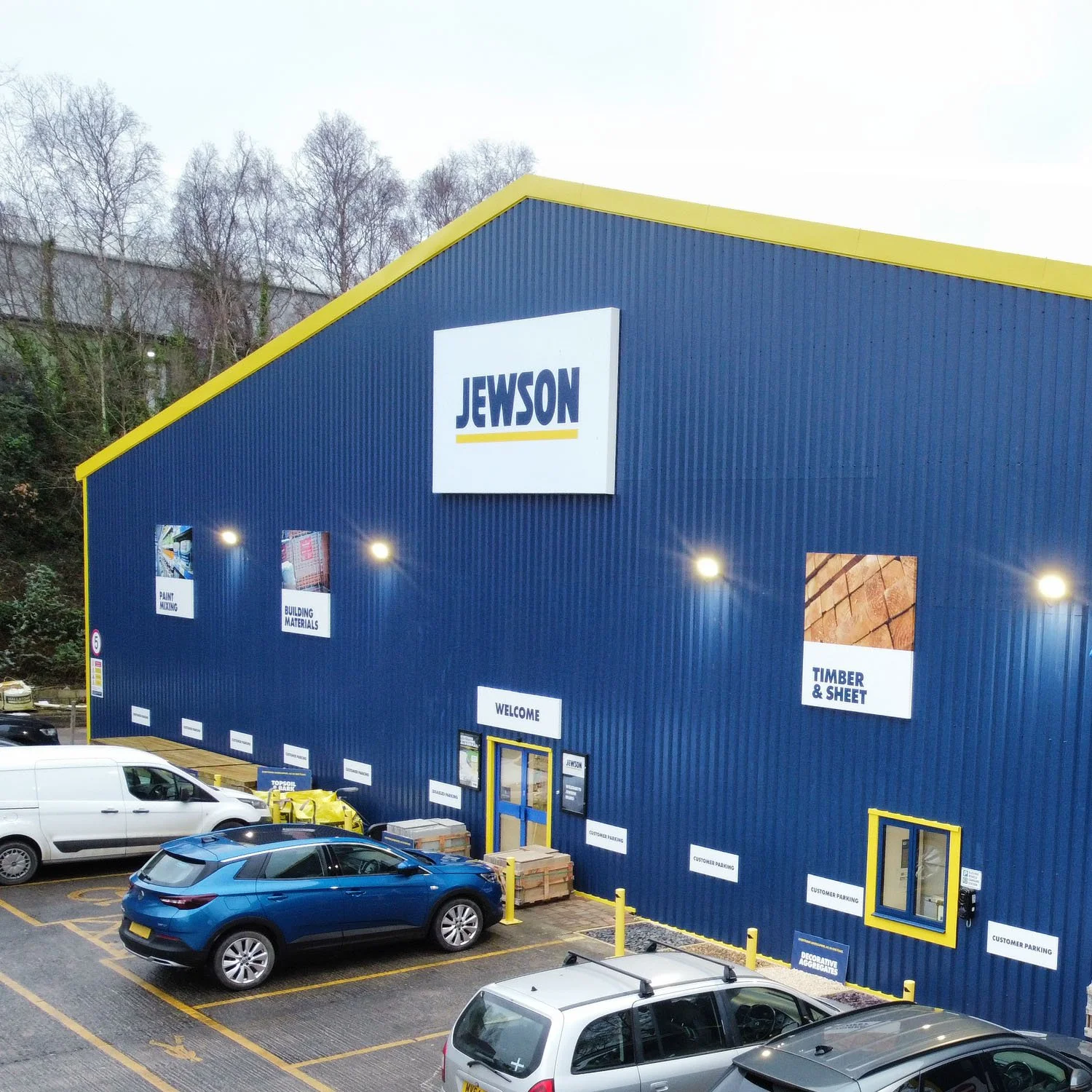 Blue building with yellow accents and a large sign that reads 'JEWSON'. Several parked cars in front, including a white van and a blue car. Signs on the building indicate building materials, timber, sheet, paint, and customer parking.