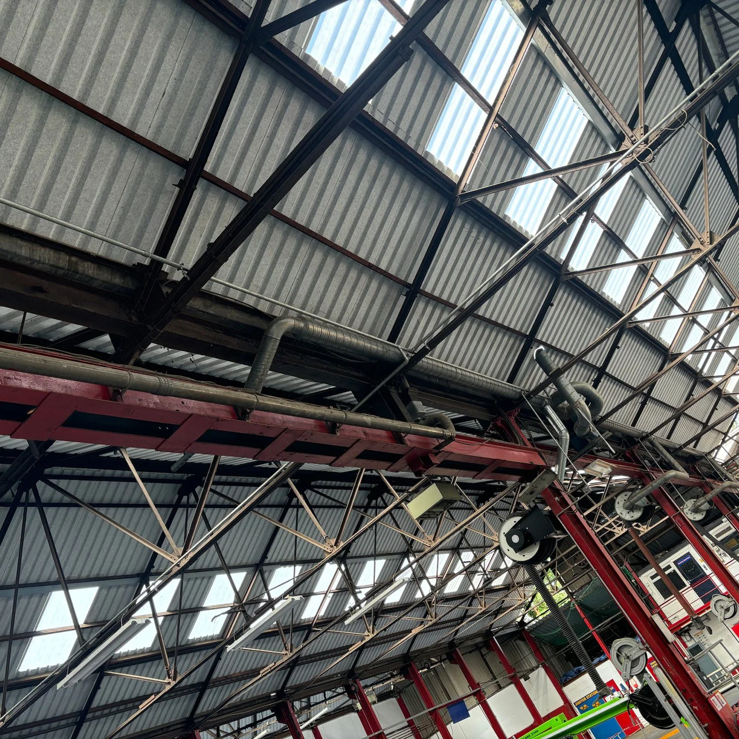 Interior of a large industrial building with a corrugated metal roof, exposed beams, and visible ducts, along with some machinery and equipment at the bottom of the image.