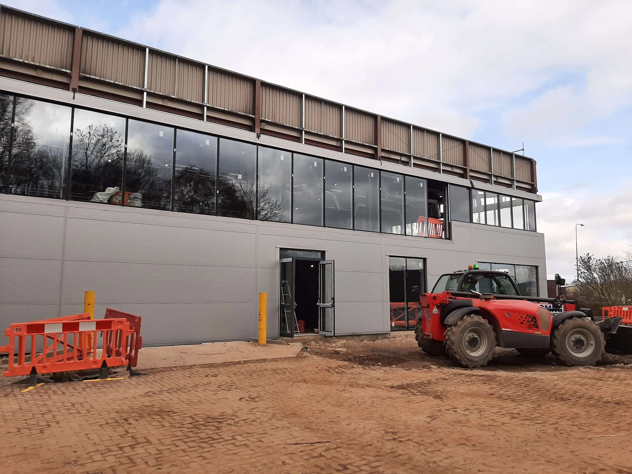 Building under construction with large glass windows, construction equipment including a red tractor, and orange safety barriers on a dirt and brick paved ground.