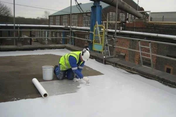 Worker in safety gear applying white waterproofing coating to a rooftop surface.