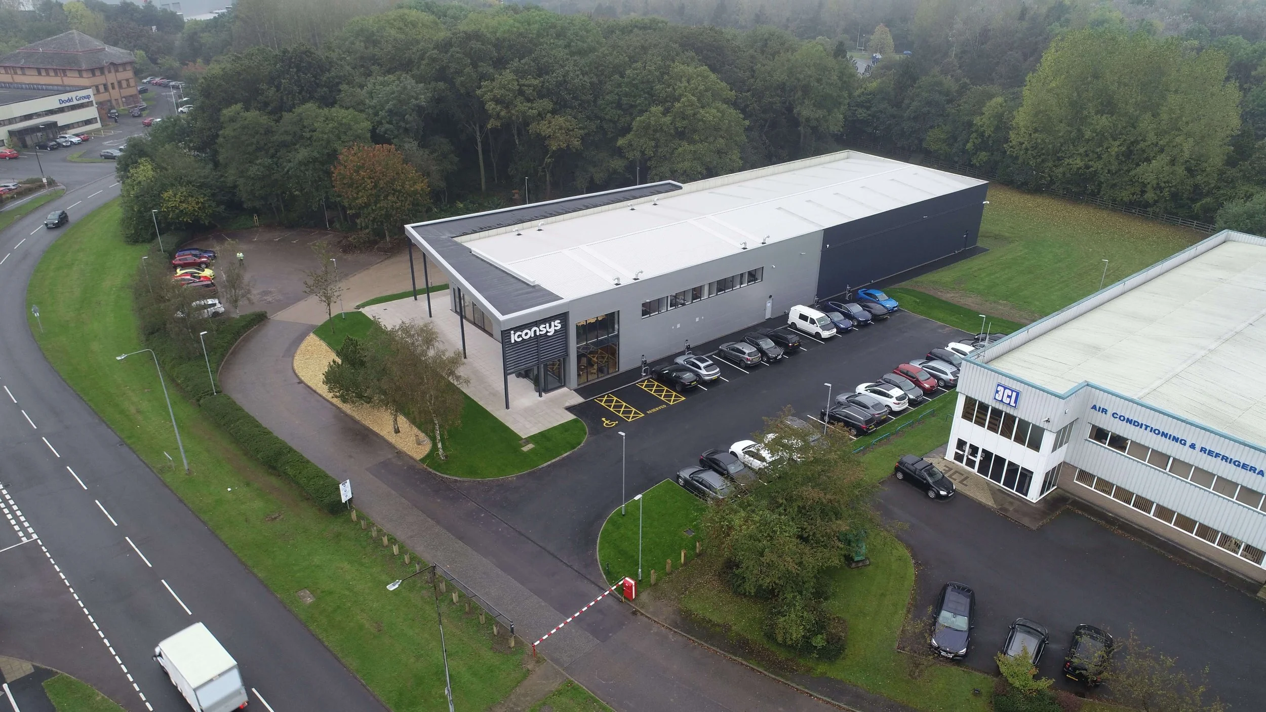 Aerial view of a commercial building with a parking lot, surrounded by trees and nearby roads.