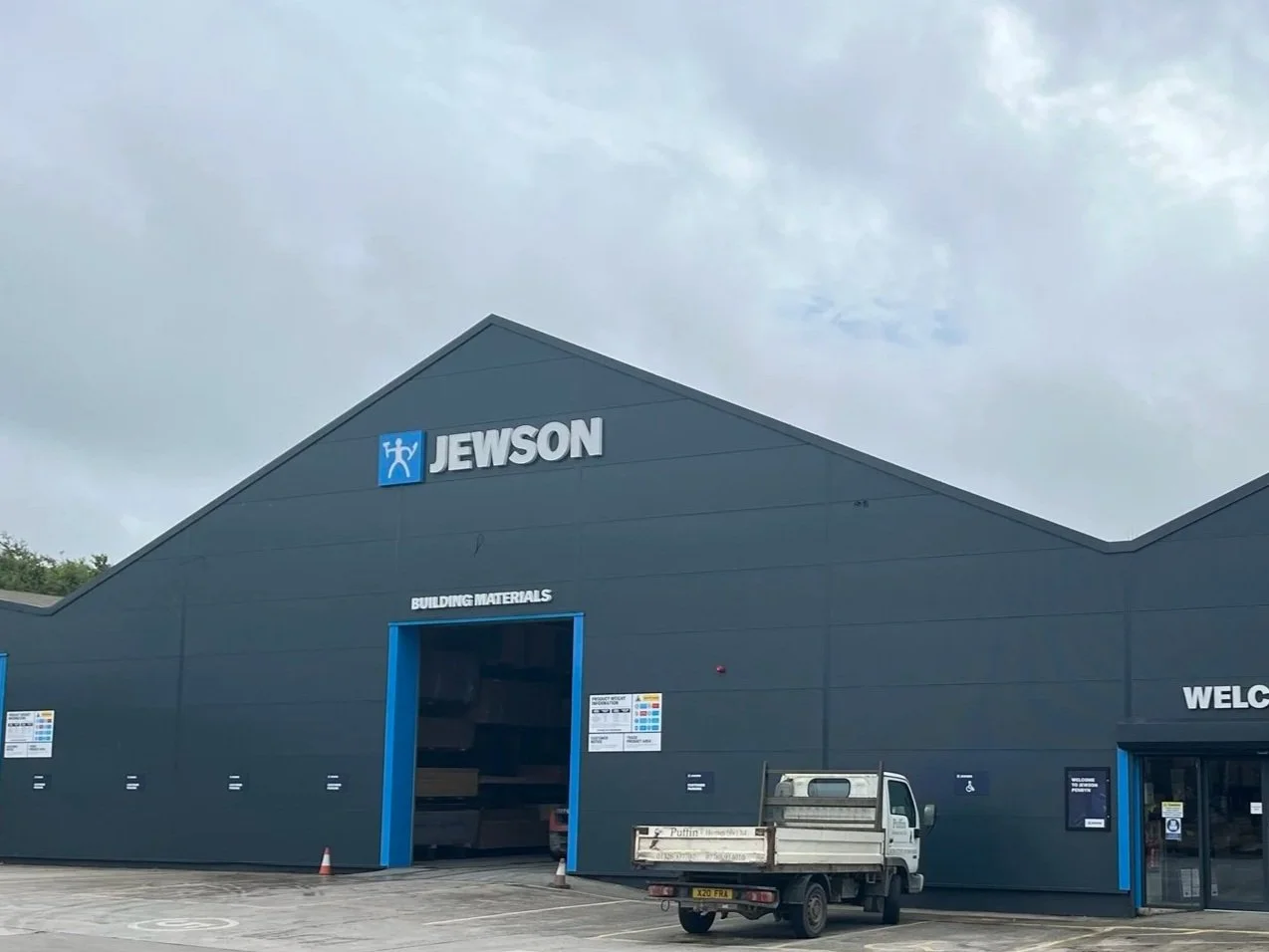Exterior of a large blue building with the sign 'JEWSON' on top, labeled 'BUILDING MATERIALS' with a truck parked outside.