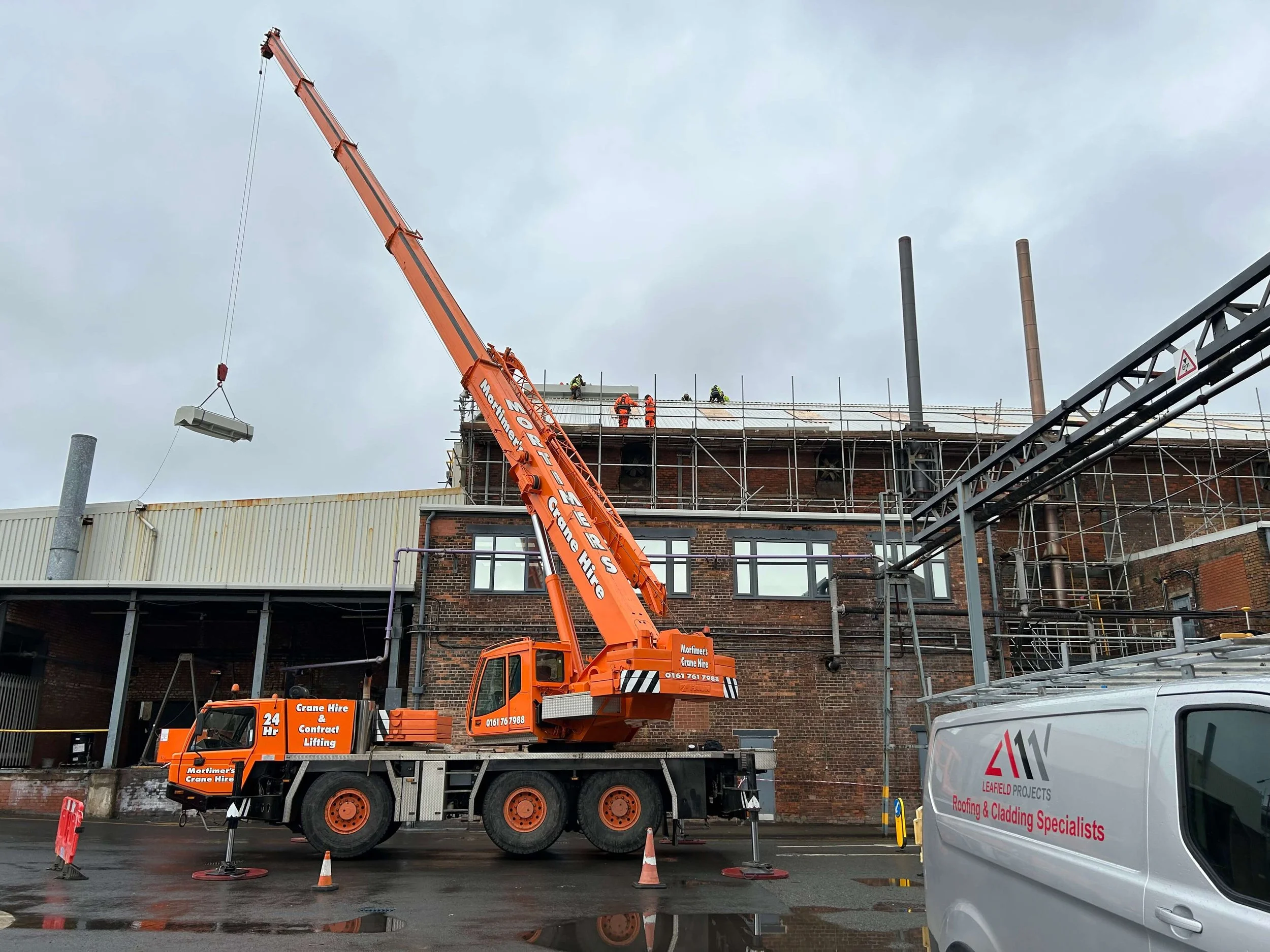 Construction workers on scaffolding working on building roof, orange crane lifting material, surrounded by construction cones, cloudy sky, nearby vehicles.