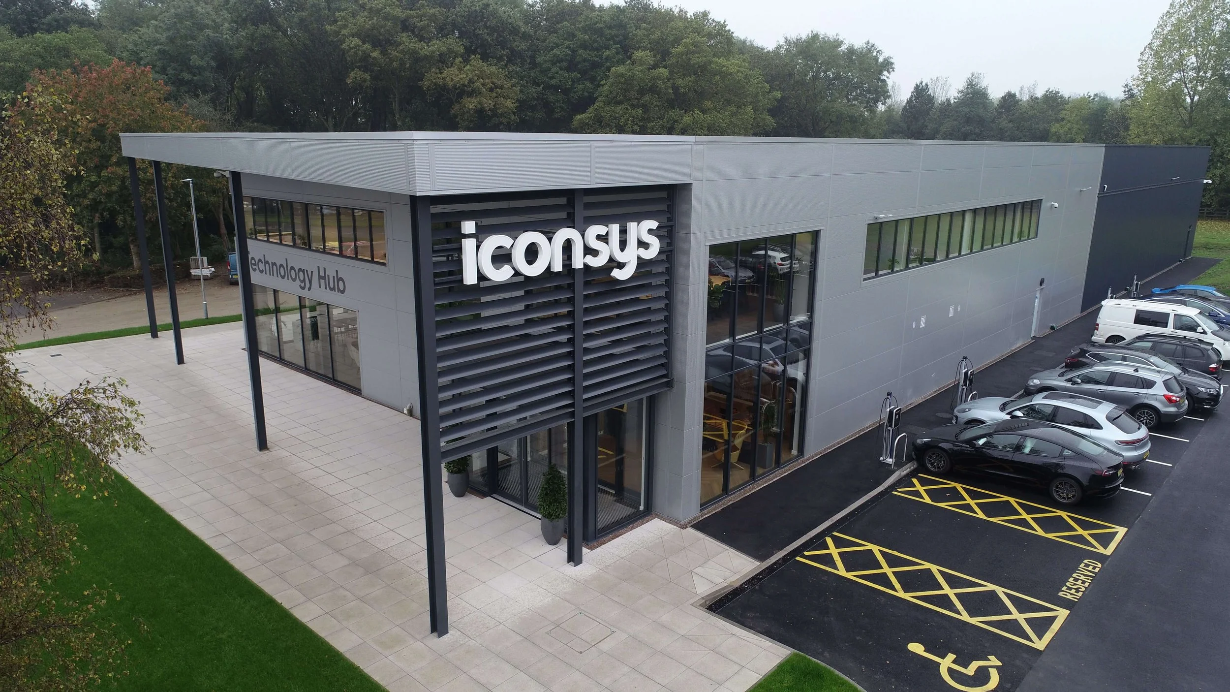 Modern two-story building with sign displaying 'iconsys' and 'technology Hub', surrounded by parking lot with several parked cars and greenery.