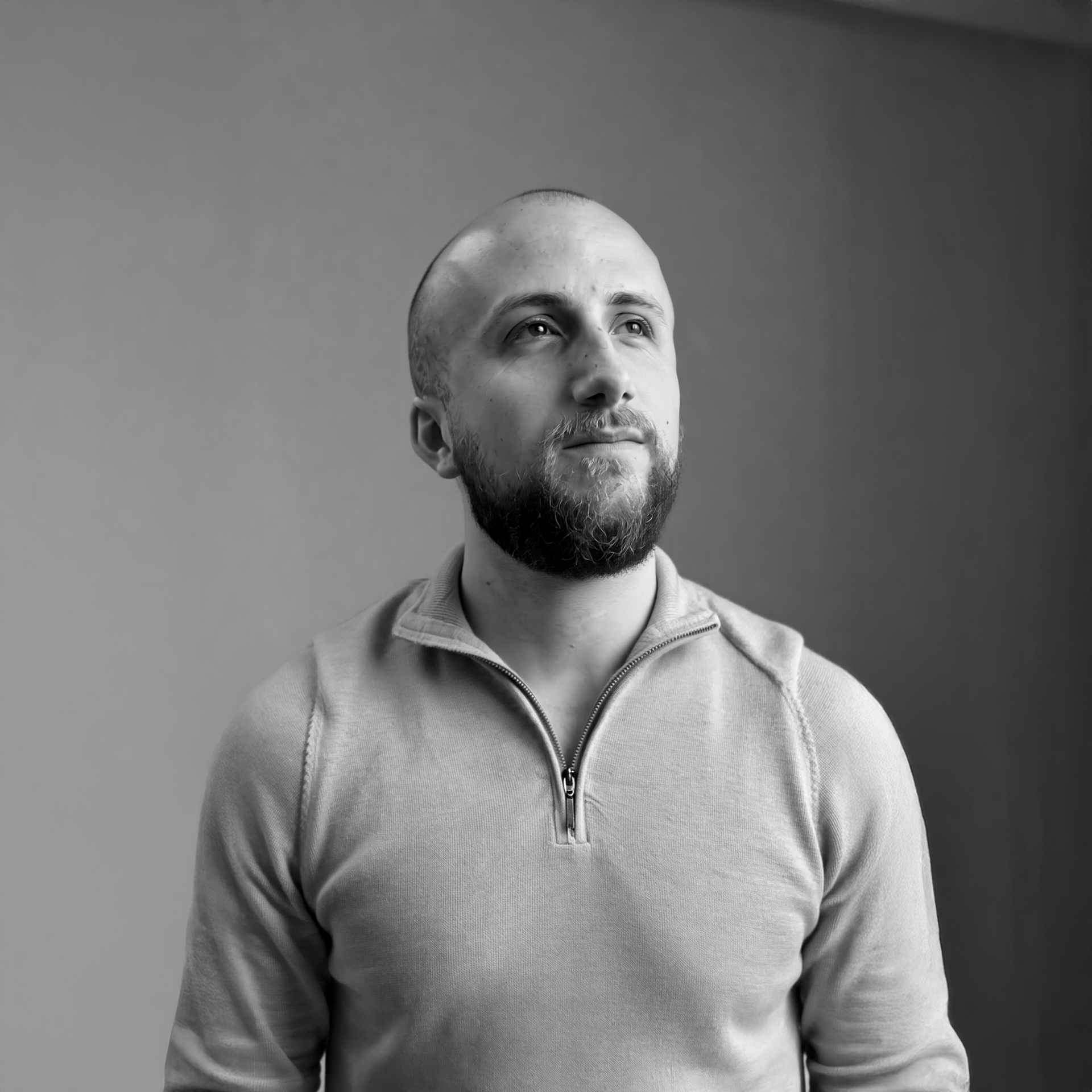 A black-and-white photo of a man with a beard and a shaved head, gazing upward with a neutral expression, wearing a zippered sweater against a plain background.