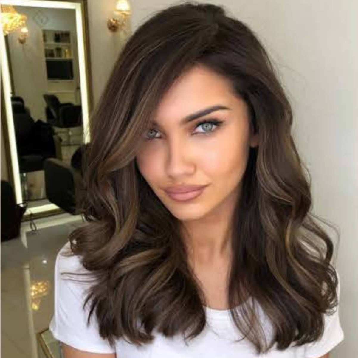 A woman with wavy brown hair and blue eyes looking at the camera in a salon with mirrors and chairs in the background.