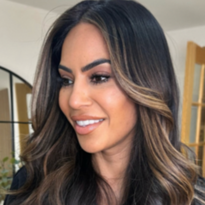 Close-up of a woman with long dark hair and highlights, smiling and looking to her left, indoors.