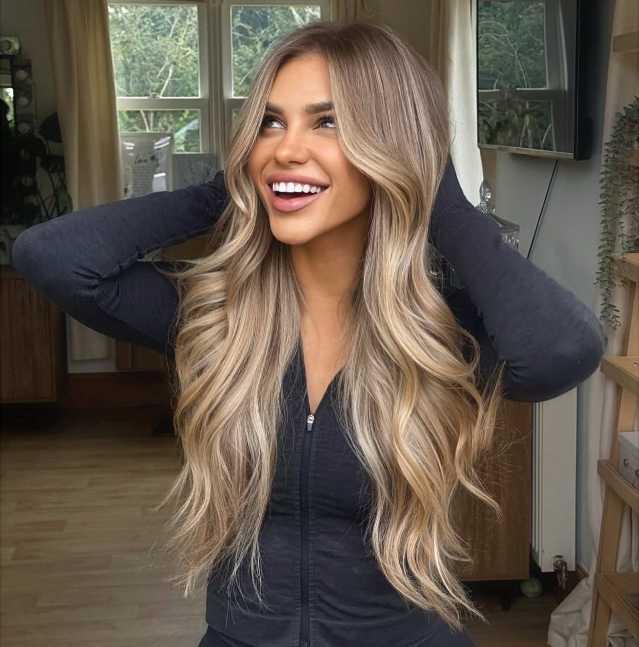 A woman with long, wavy blonde hair extensions smiling and holding her hands behind her head indoors.