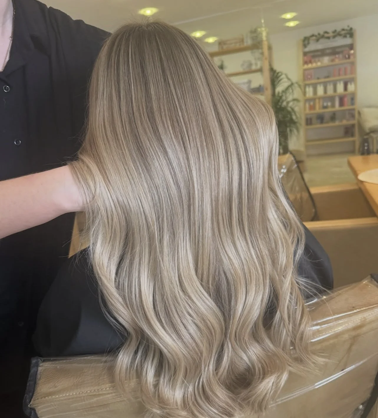 A person with long, wavy, blonde hair sitting in a salon chair, with a stylist in black standing nearby.