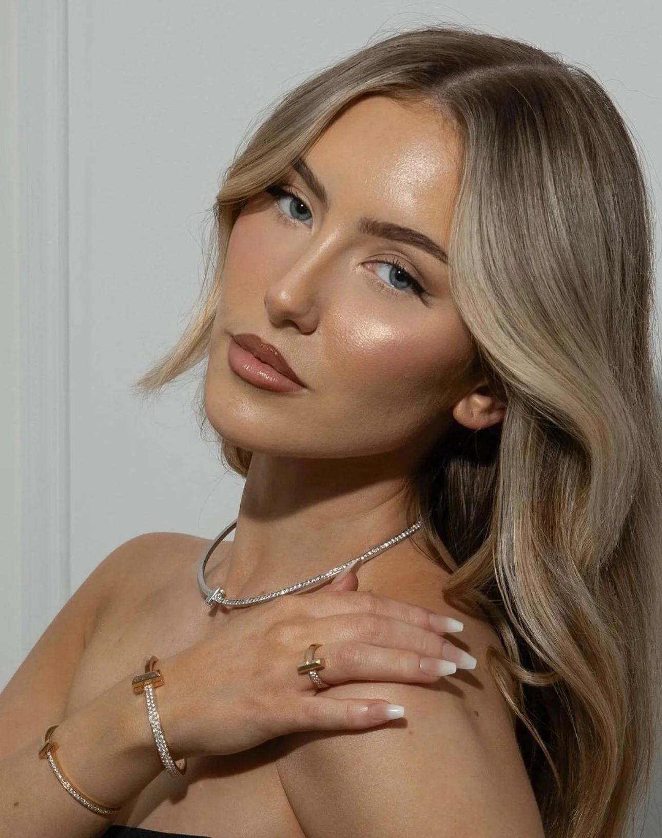 A woman with wavy blonde hair and blue eyes wearing jewelry, including a silver necklace, rings, and bracelets.