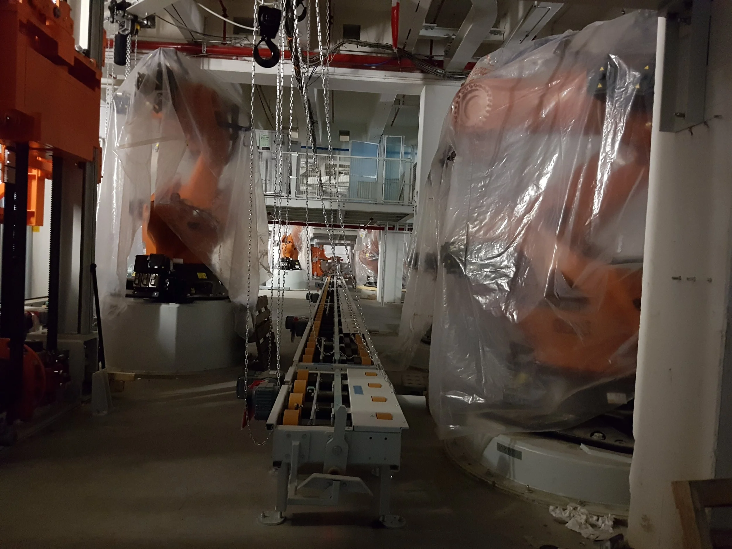 Industrial robotic arms covered in plastic, working in a manufacturing or assembly facility with an overhead conveyor system.