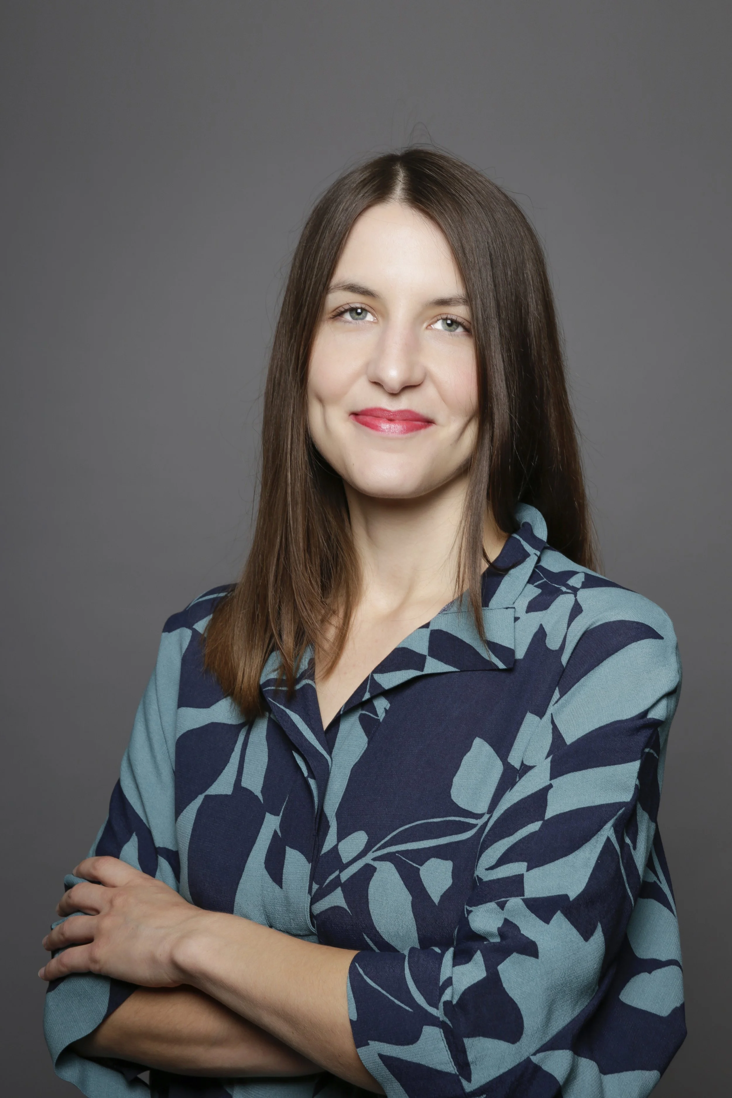 A woman with straight brown hair and fair skin, wearing a blue patterned blouse, standing with arms crossed in front of a gray background, smiling softly.