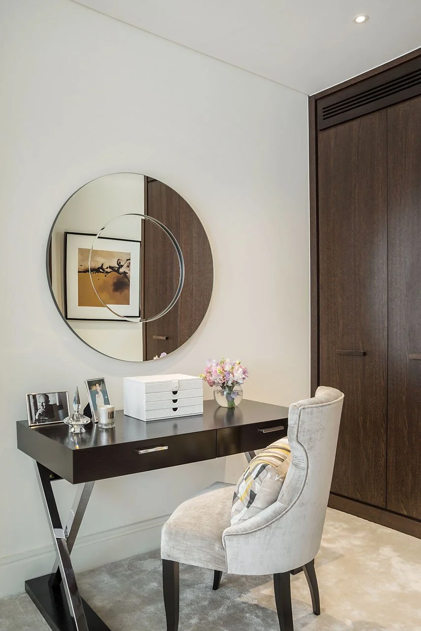 A modern home office space with a dark wooden desk, a round mirror on the wall, and a white upholstered chair with a plaid pillow. There are framed photos, a flower vase, and a white storage box on the desk.