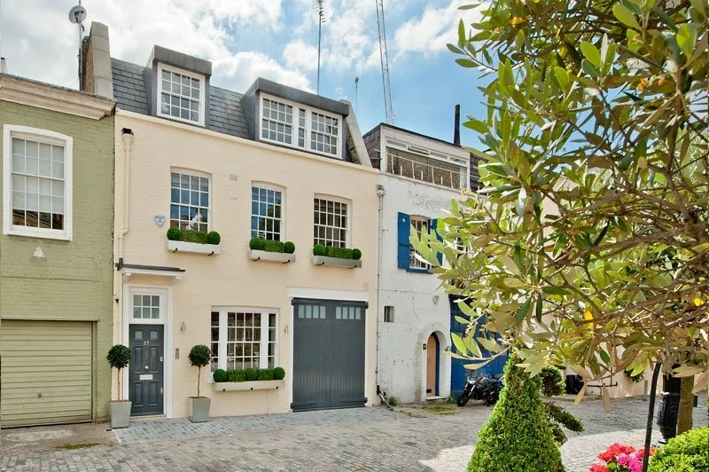 Colorful row of European-style townhouses with potted plants and cobblestone street, trees, and sky in the background.