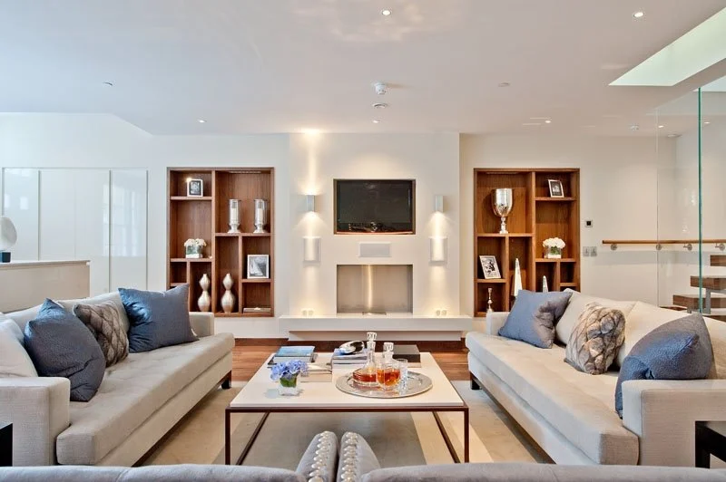 Living room with two beige sofas, a white coffee table with drinks and decor, built-in shelves with decorations, a wall-mounted TV, and modern lighting.