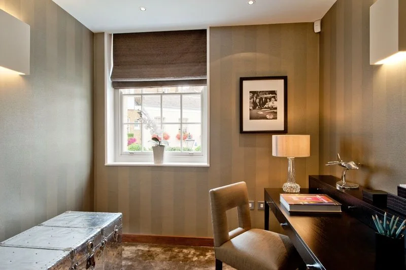 A cozy home office with a window, desk, chair, table lamp, framed black-and-white photo, and decorative items, illuminated by warm lighting.