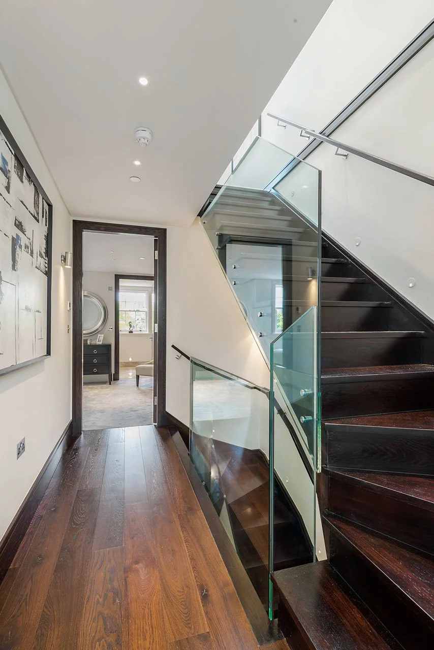 Interior view of a modern staircase with glass railing leading upstairs, wooden flooring, and a doorway to a room with a window and a dresser.