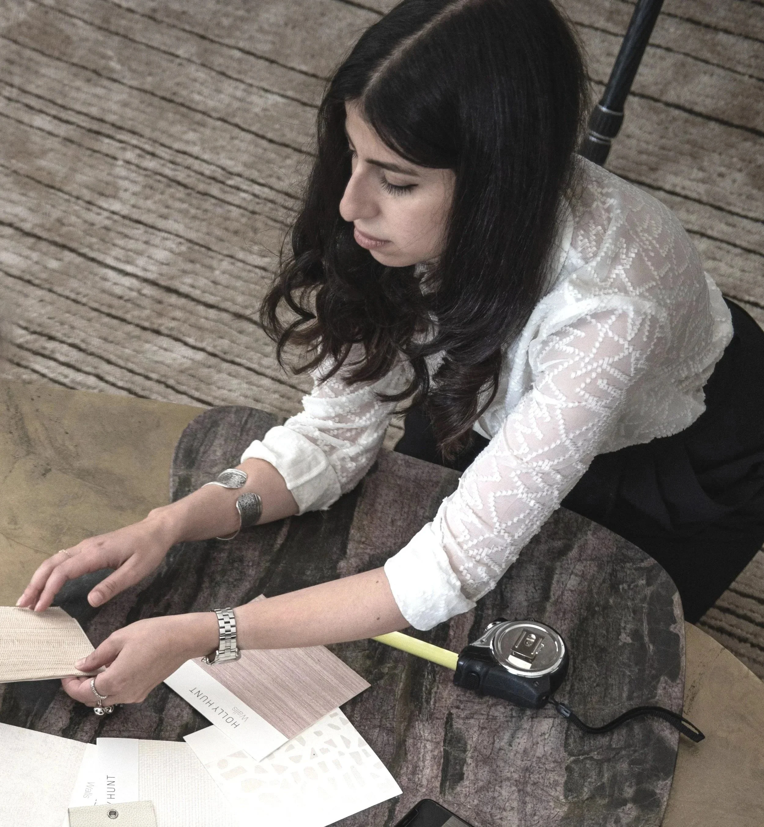 A woman with long dark curly hair, wearing a white lace blouse and black pants, is sitting at a table and examining wood samples. The table has fabric and paper samples, a digital camera, and a smartphone.