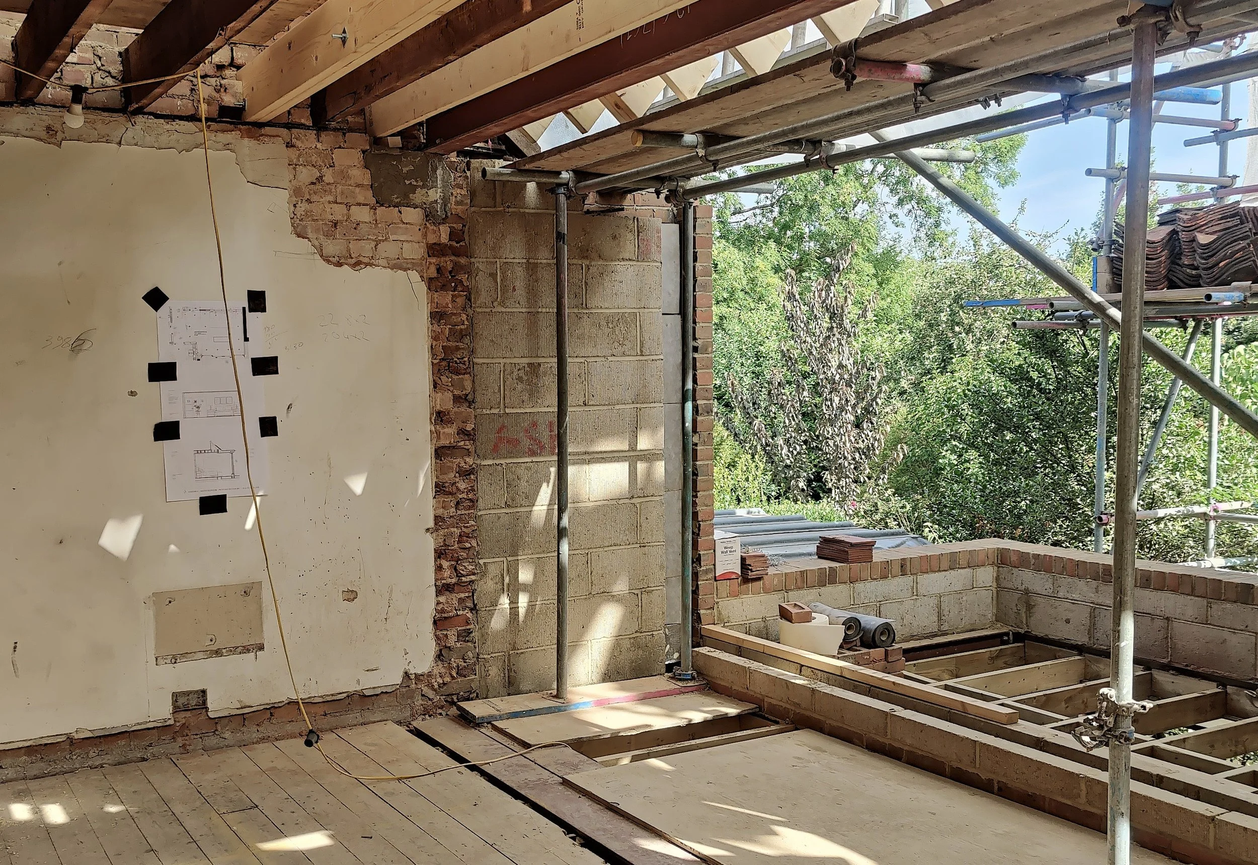 Construction site with partially built brick walls, scaffolding, and scaffolding planks. The background shows trees and a partly cloudy sky. Site drawings and construction details are taped to the wall