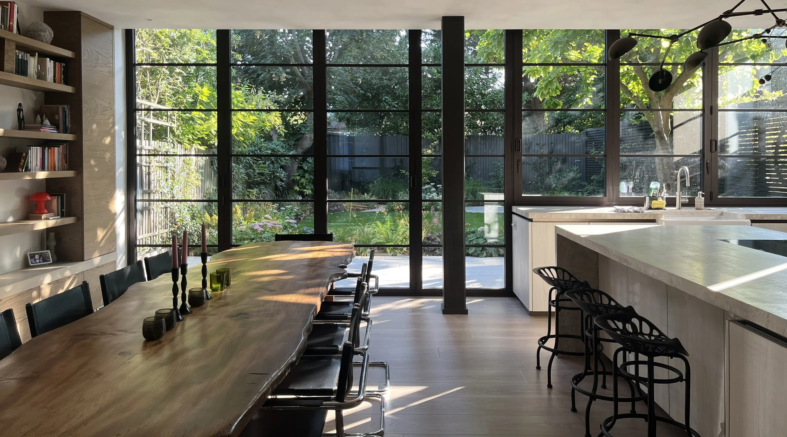 Modern kitchen open plan and dining area with large glass bifolding doors leading to a lush garden, a waney edge wooden dining table with candles, vintage mart stam cantilever chairs, kitchen with taj mahal worktop and butler sink