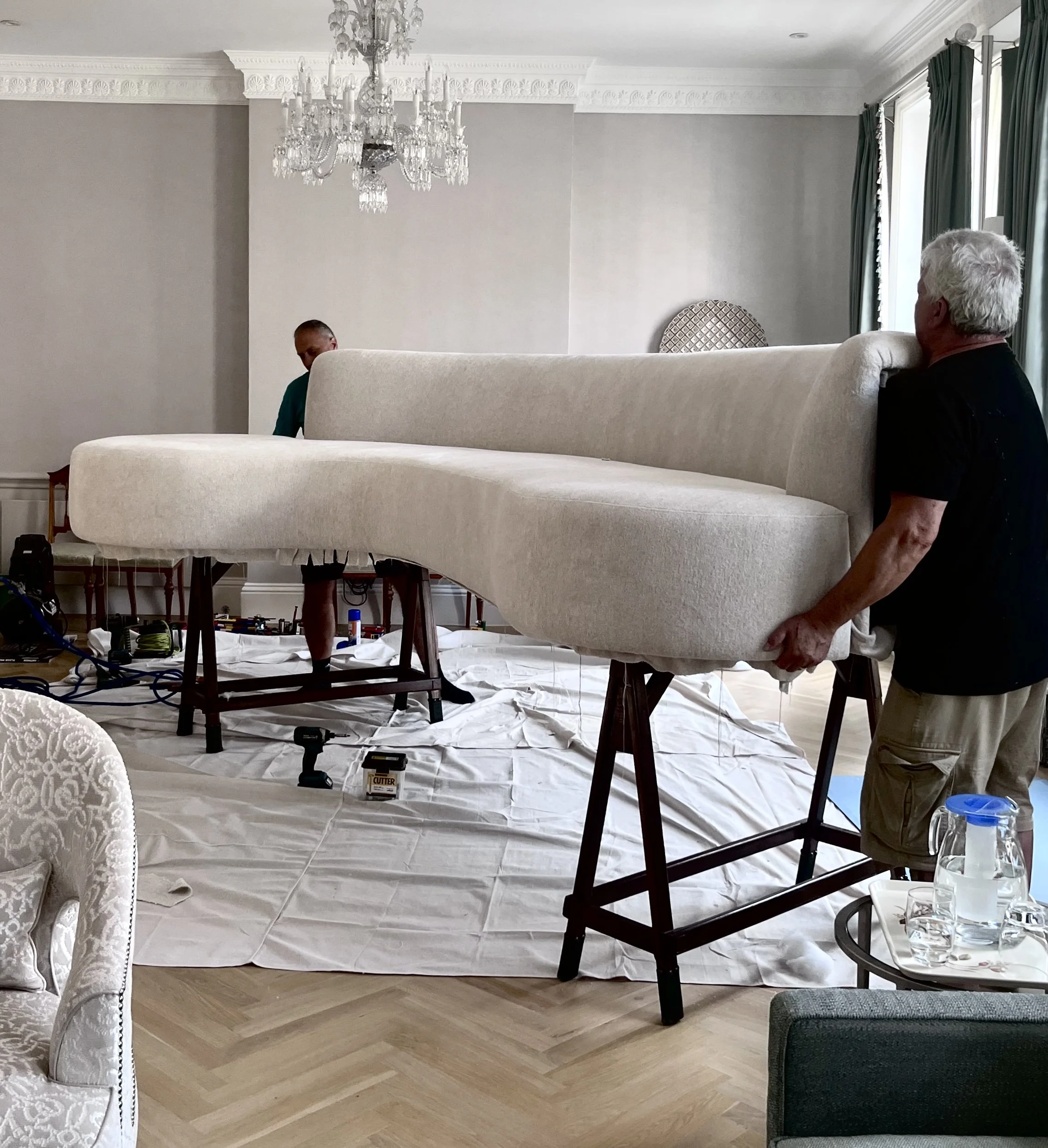 Two men are making large curved cream-colored sofa in situ in a room with light walls, a chandelier, and large windows with teal curtains. The sofa is on supports and is being carried by one man, while a second man is partially visible behind it.