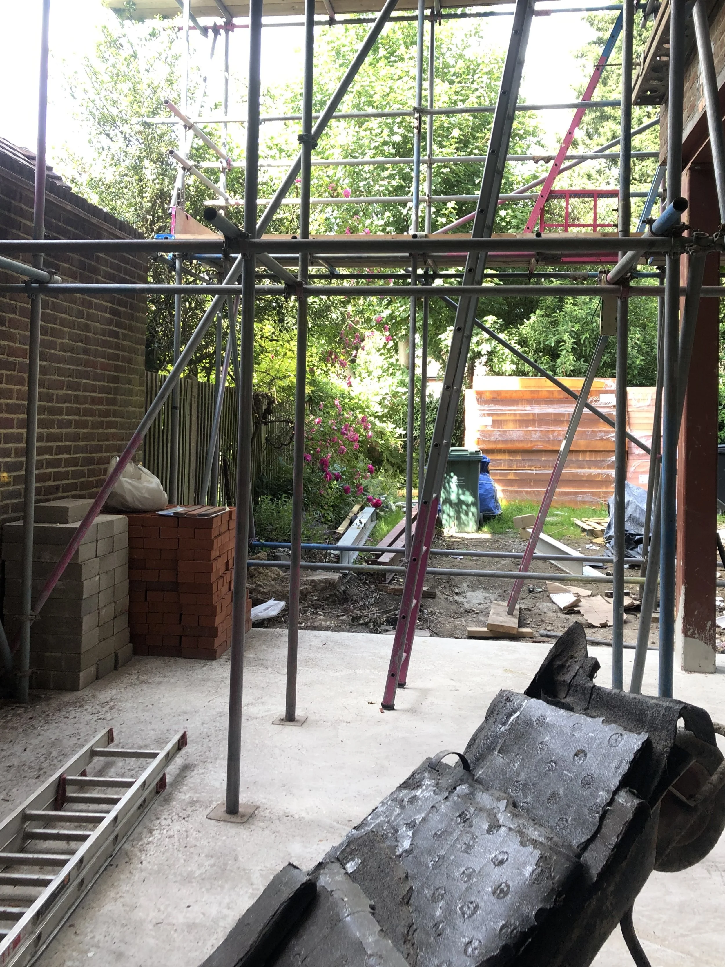 Construction site with metal scaffolding, ladders, bricks, and construction materials, with a partially completed porch or building extension, surrounded by greenery and trees.