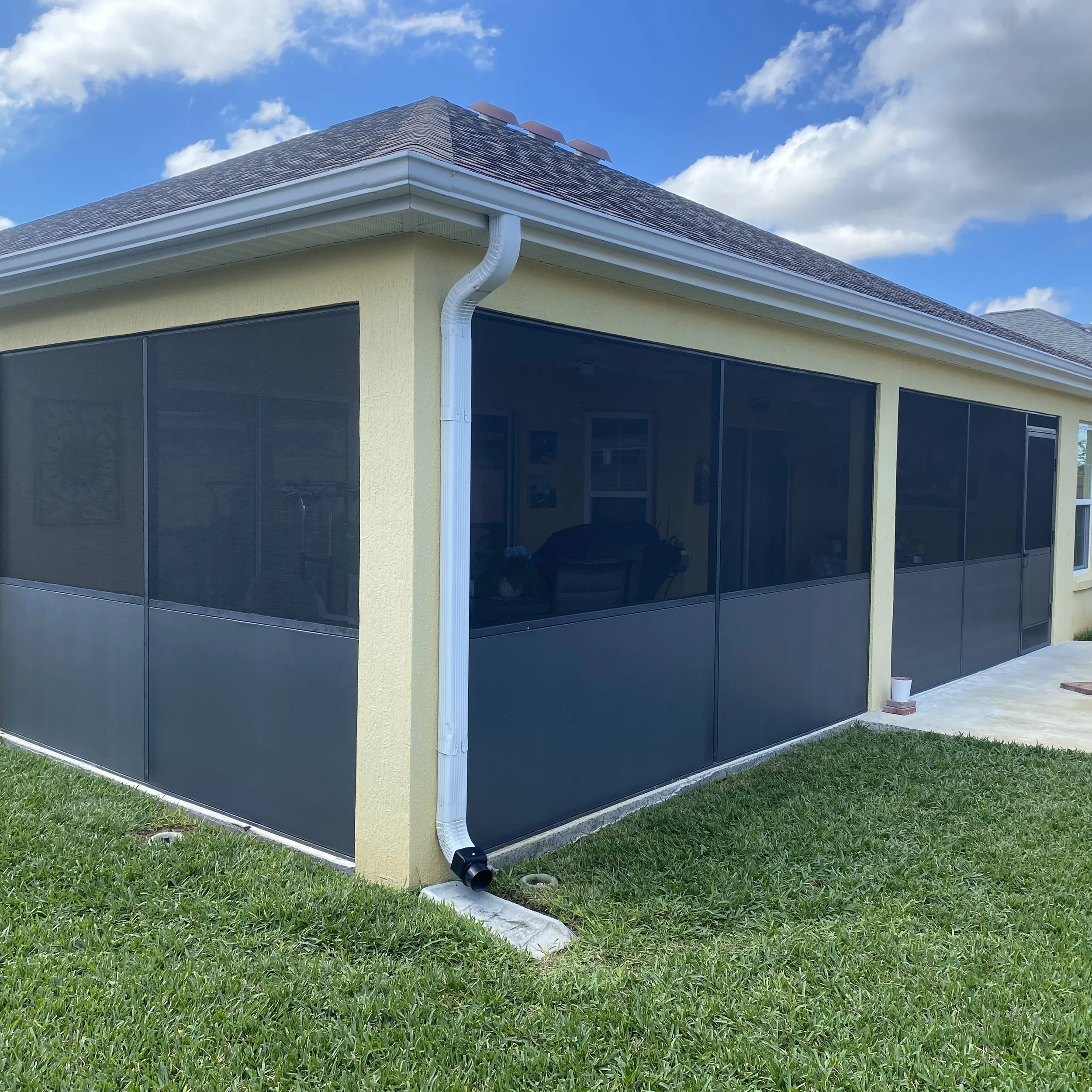 Screened-in backyard patio with a yellow house, a gray roof, a white gutter system, and a patio with a potted plant on a concrete surface. Green grass surrounds the area.