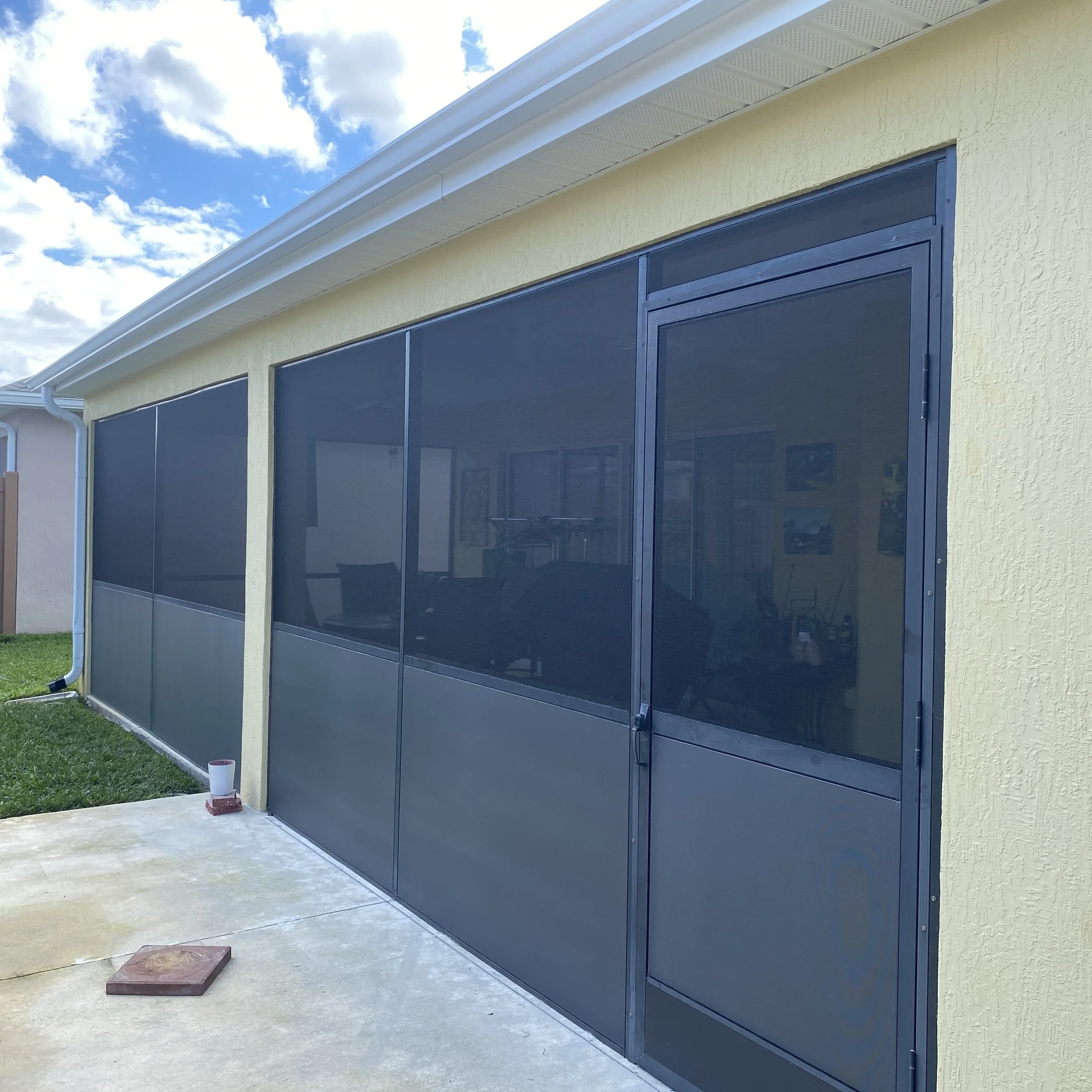 Screened porch with black mesh panels on a yellow house, concrete floor, and blue sky with clouds.