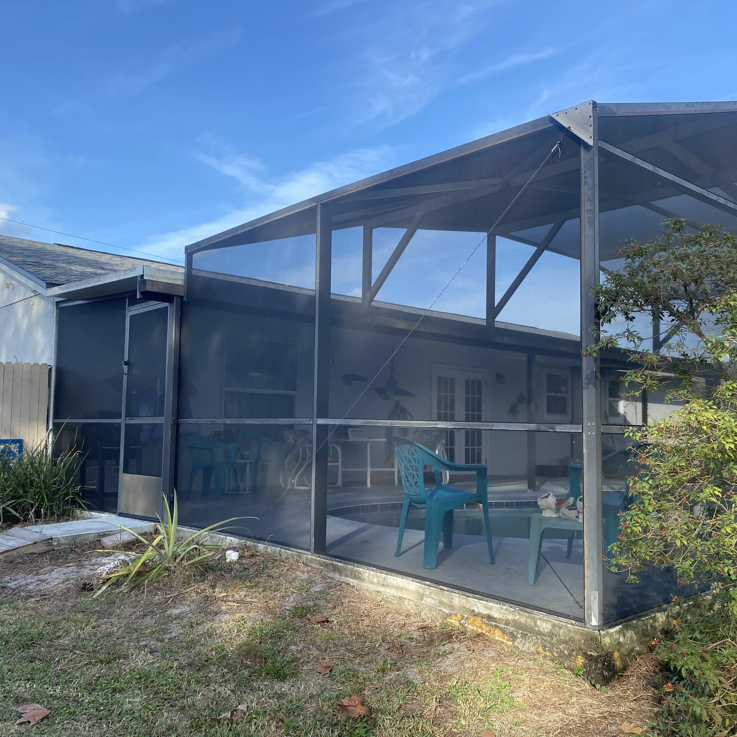 Screened porch with outdoor furniture and a small pool, in a backyard with grass and plants, under a blue sky with some clouds.