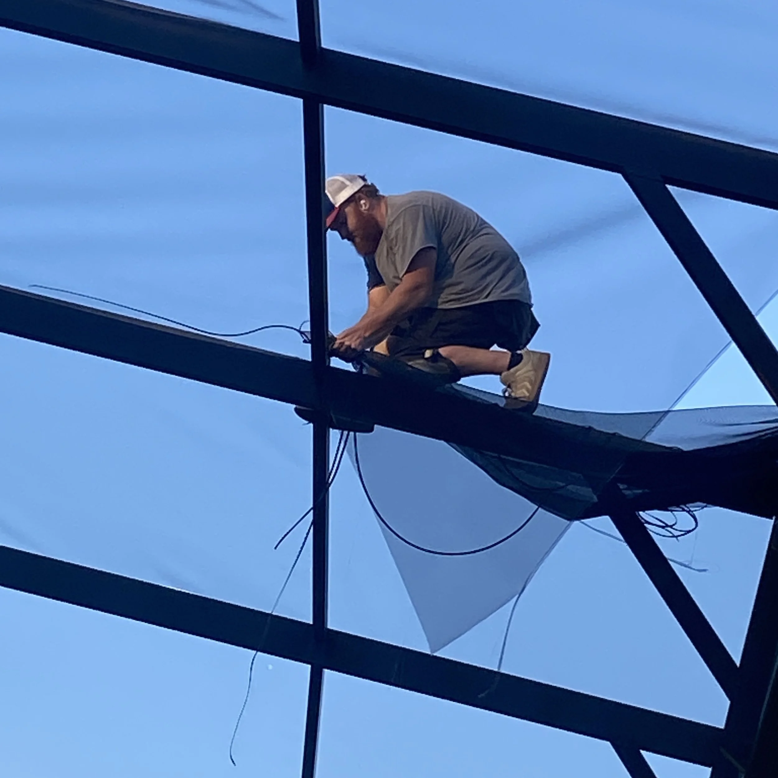 A man working on the exterior of a glass building, crouched on a platform, wearing a cap and casual clothes.