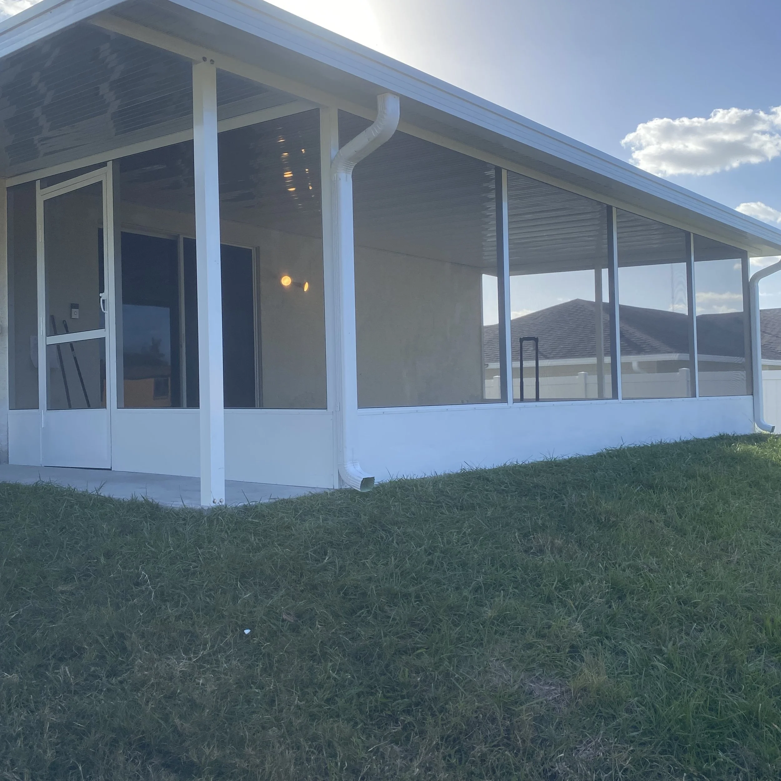 View of a house's screened-in porch with glass sliding door, white frame, and green yard in sunlight.