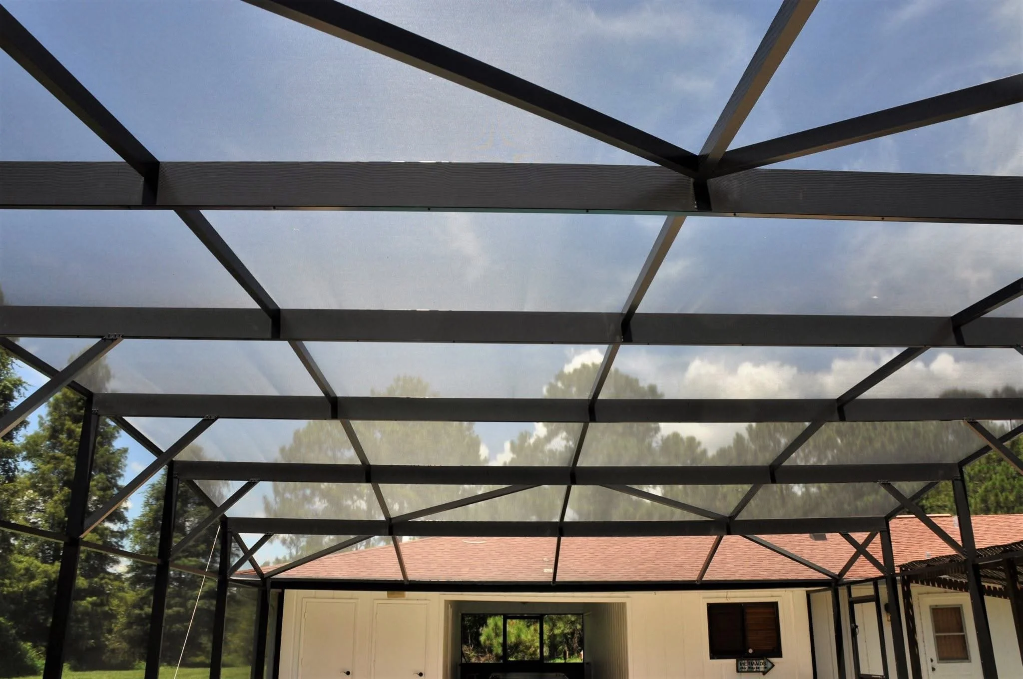 View of a screened porch with a metal frame roof, attached to a house with white walls and a red roof, surrounded by green trees and a blue sky with some clouds.