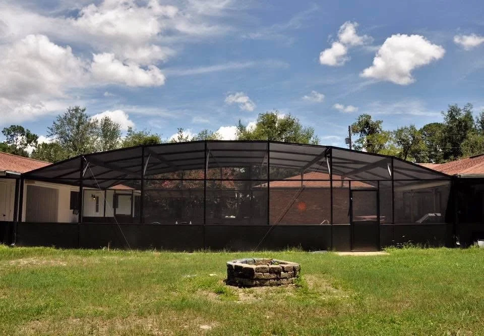 A screened-in patio or porch with a black metal frame and a translucent roof, attached to a beige house in a backyard with green grass and a fire pit in the foreground. The sky is partly cloudy with some blue showing.